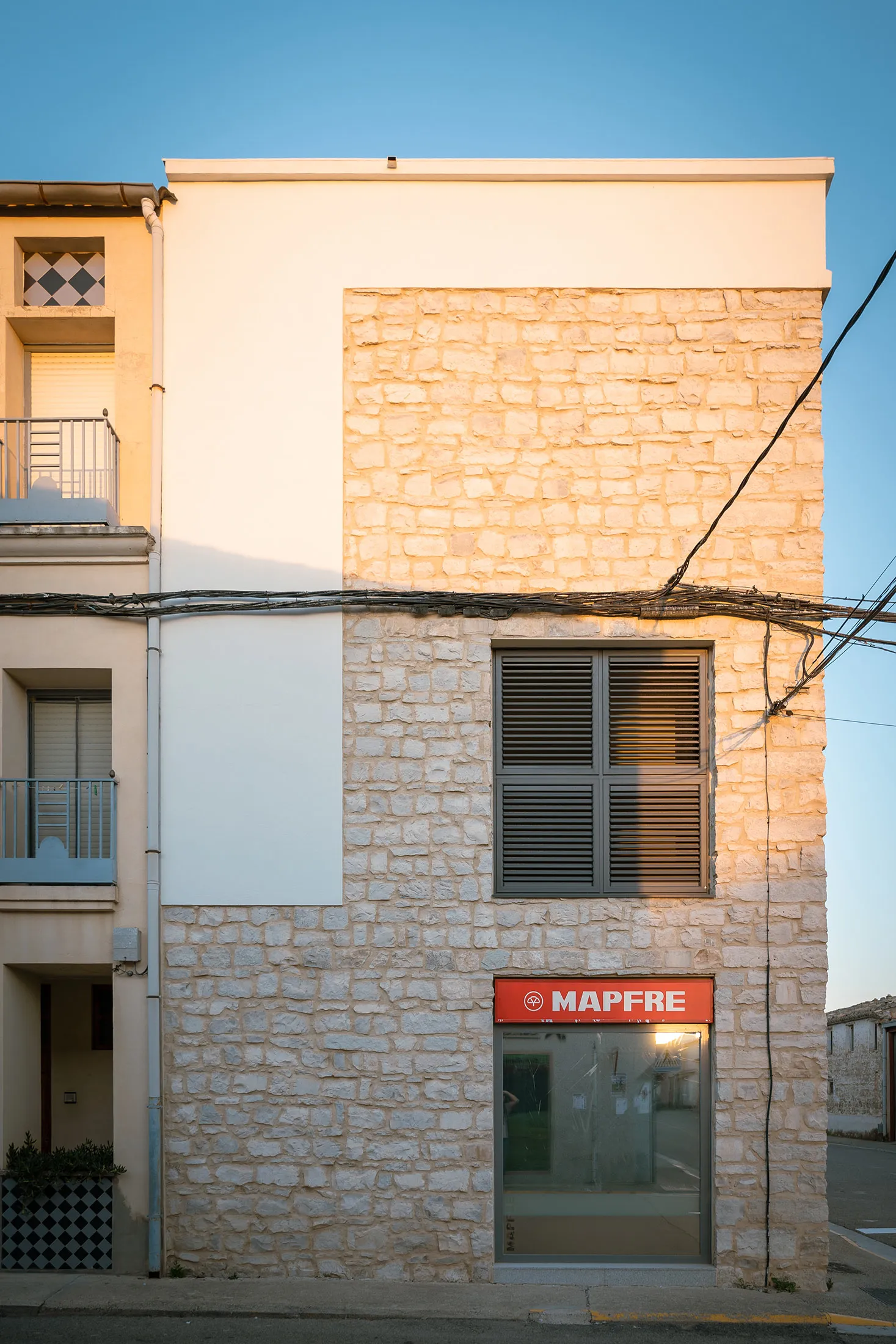 Composición de fachada combinando paños de piedra y enfoscado blanco, con un gran ventanal y rótulo